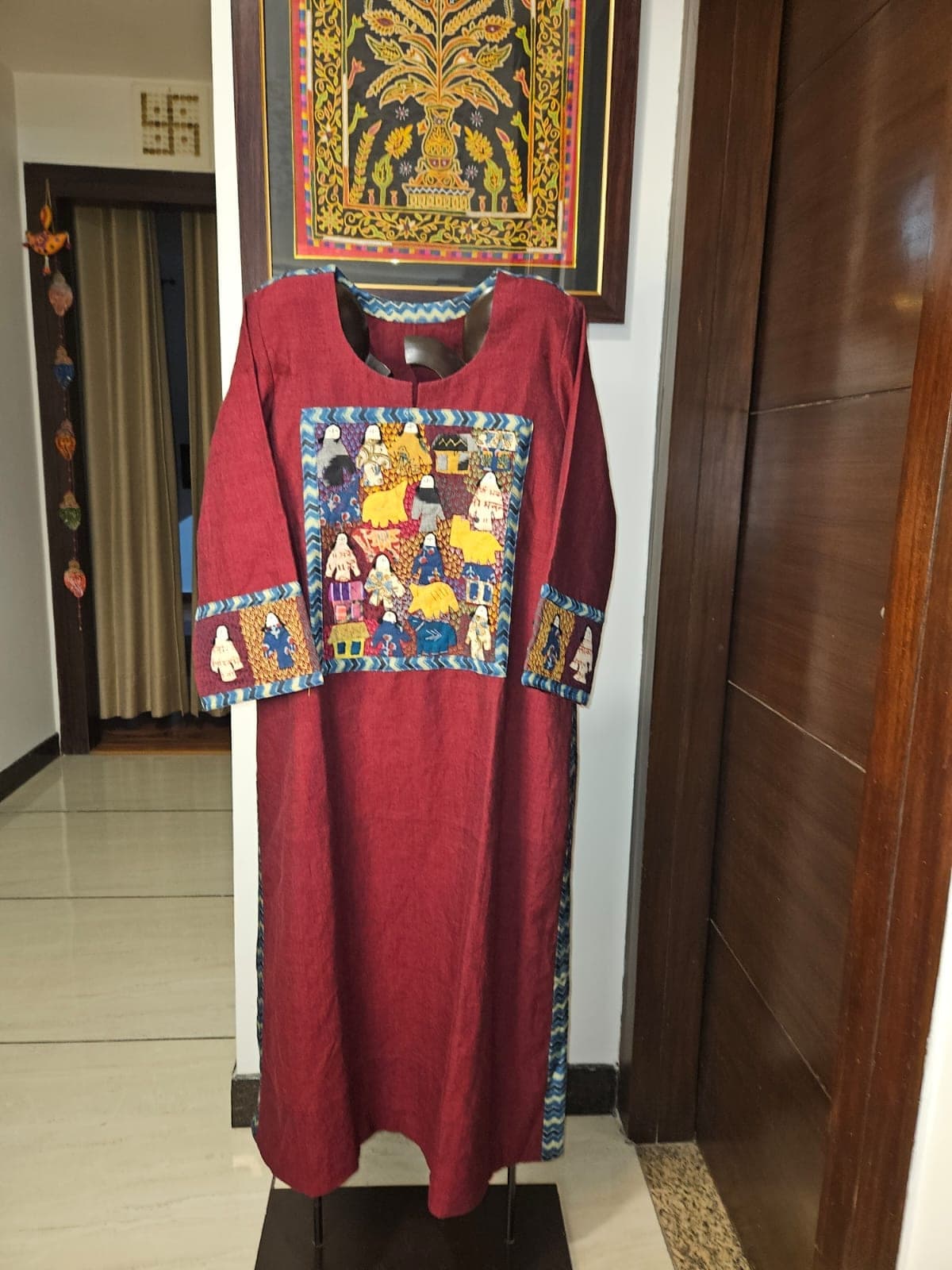 Maroon kurti with a patchwork panel of people, animals and houses, and matching cuff panels, on a stand in a hallway